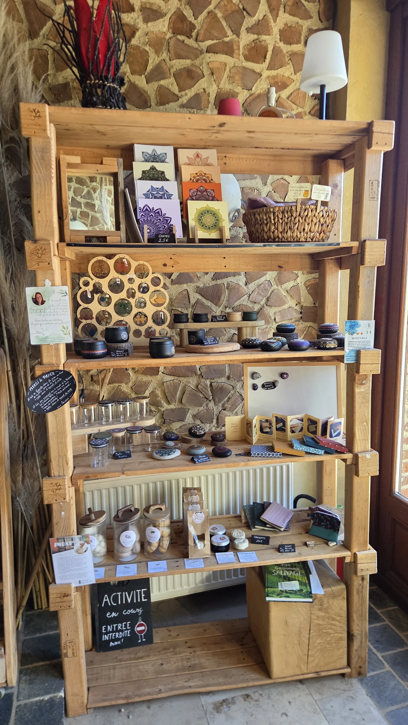 Étagère en bois de l'épicerie chargée d'artisanat : livrets de mandalas, savons, pots, créations diverses, mur en pierres apparentes derrière