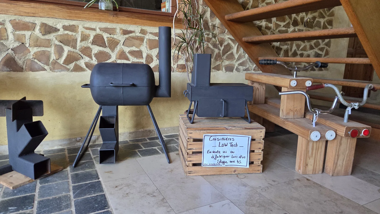 Démonstration low-tech : un poêle fabriqué dans une bombonne métallique posée sur des cales en bois à côté d'un autre poêle en forme d'animal, un banc avec une caisse en bois et un panneau d'explication écrit à la main