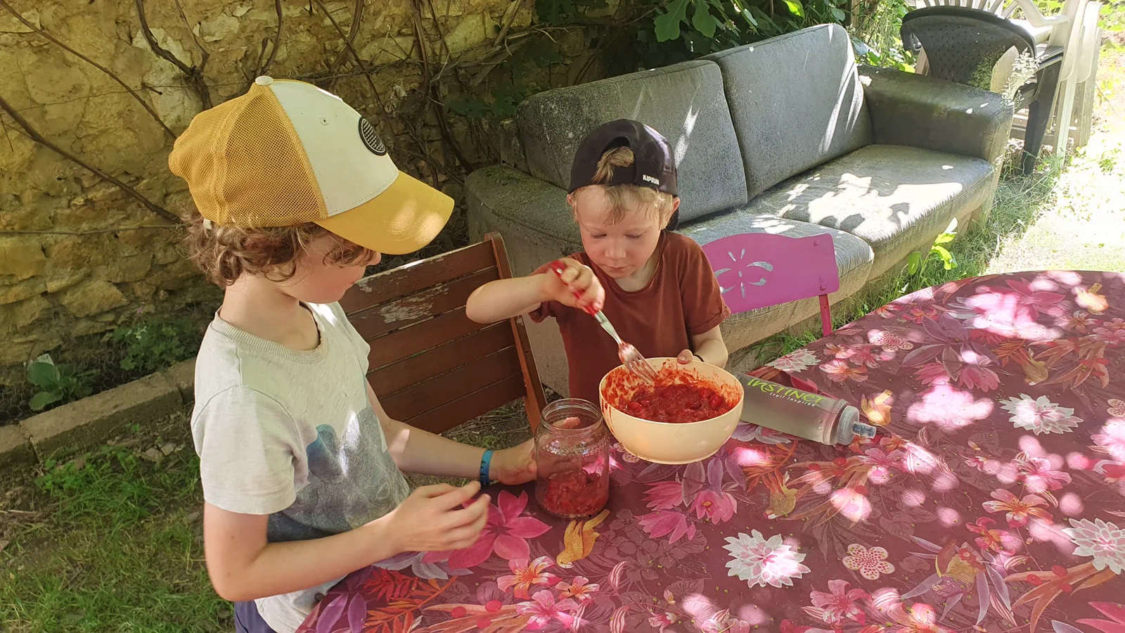 Colin et Léon assis à une table de jardin sous une nappe à fleurs, en train de touiller un grand saladier de confiture de fruits rouges, canapé d'extérieur derrière