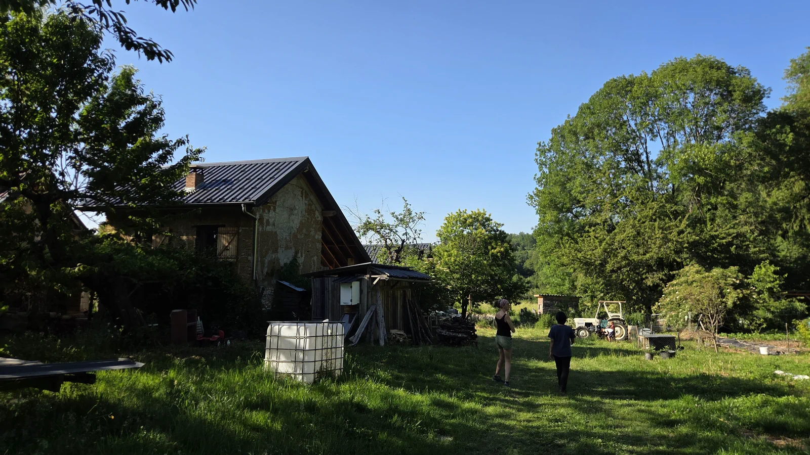 La maison de Marie-Laure, ancienne bâtisse en pierre au toit rénové, dans un pré en pente, deux personnes qui marchent vers le potager et un tracteur au fond