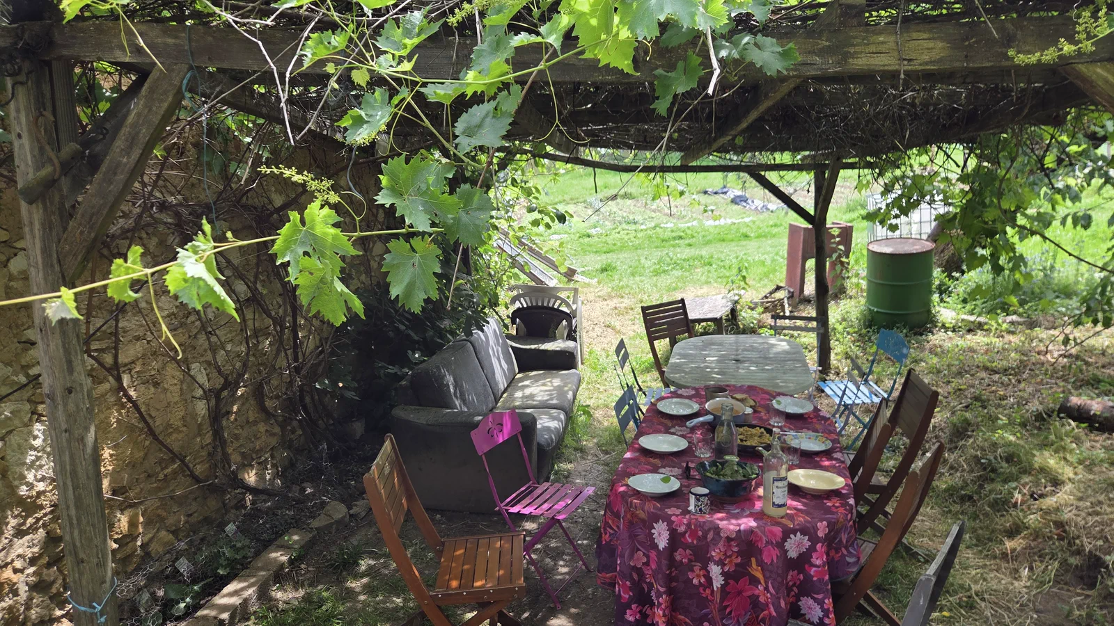 Coin repas en plein air sous une pergola de vigne, longue table couverte d'une nappe à fleurs et de vaisselle, canapé et chaises dépareillées, ouverture sur le pré derrière