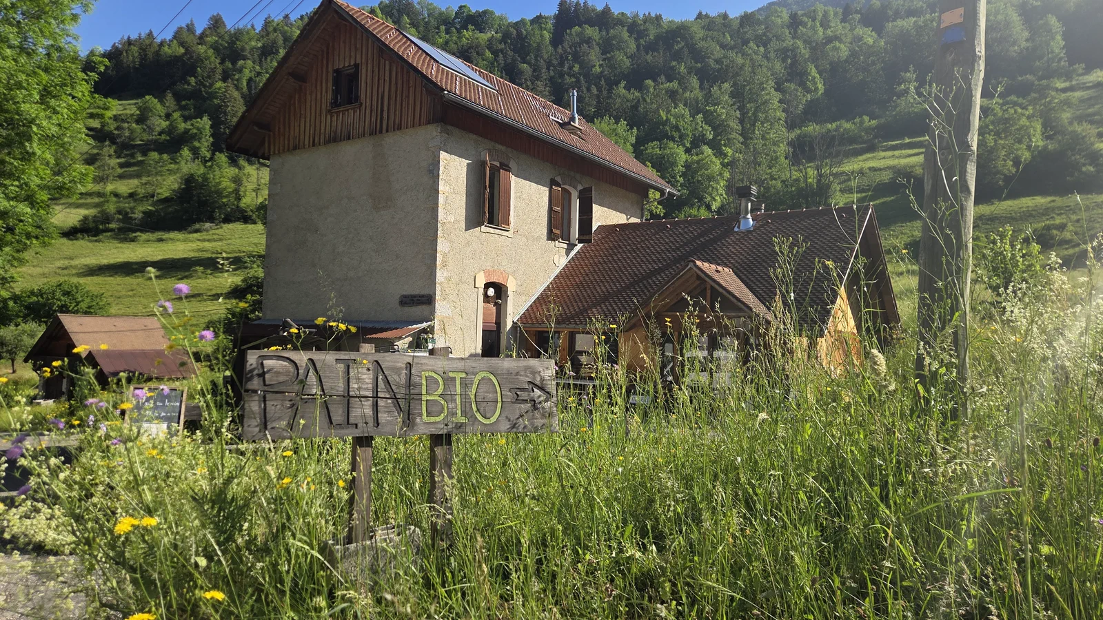 L'ancienne école en pierre devenue fournil, sur un versant verdoyant de Chartreuse, avec au premier plan un panneau en bois « PAIN BIO » dans les herbes folles et les fleurs