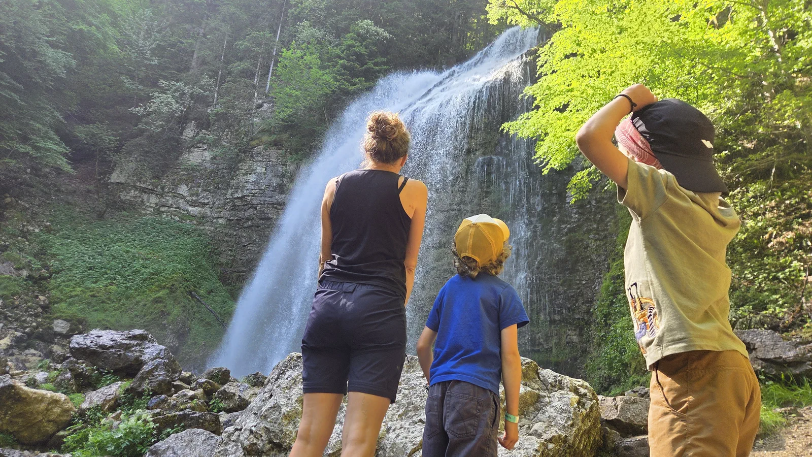 La famille de dos face à la grande cascade du Cirque de Saint-Même, falaise et forêt verdoyante autour, rochers au premier plan