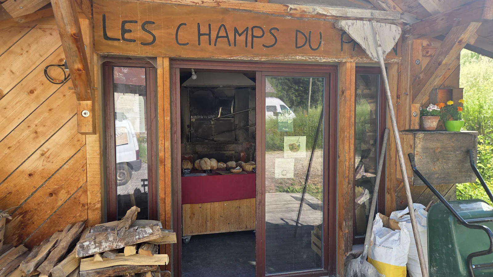 Entrée vitrée du fournil avec l'enseigne « Les Champs du Pain » gravée dans le bois, pains posés sur une table à l'intérieur, bûches empilées et sac de farine Moulin Marion à droite