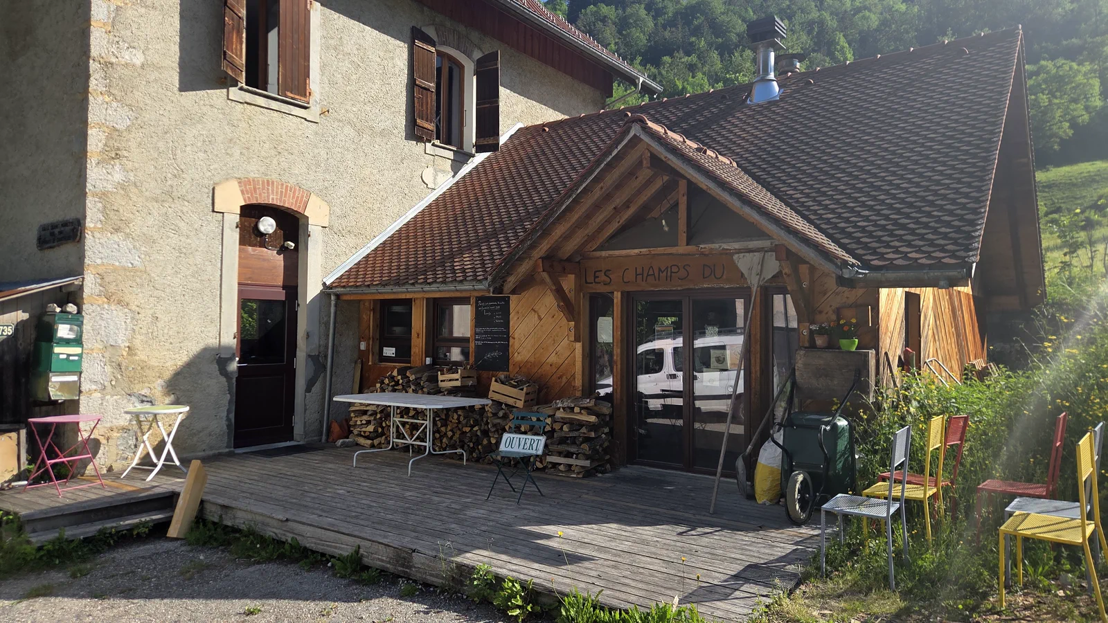 Façade du fournil installé dans l'ancienne école en pierre, auvent en bois avec l'enseigne « Les Champs du Pain », terrasse avec tables et chaises dépareillées, bûches empilées devant l'entrée vitrée