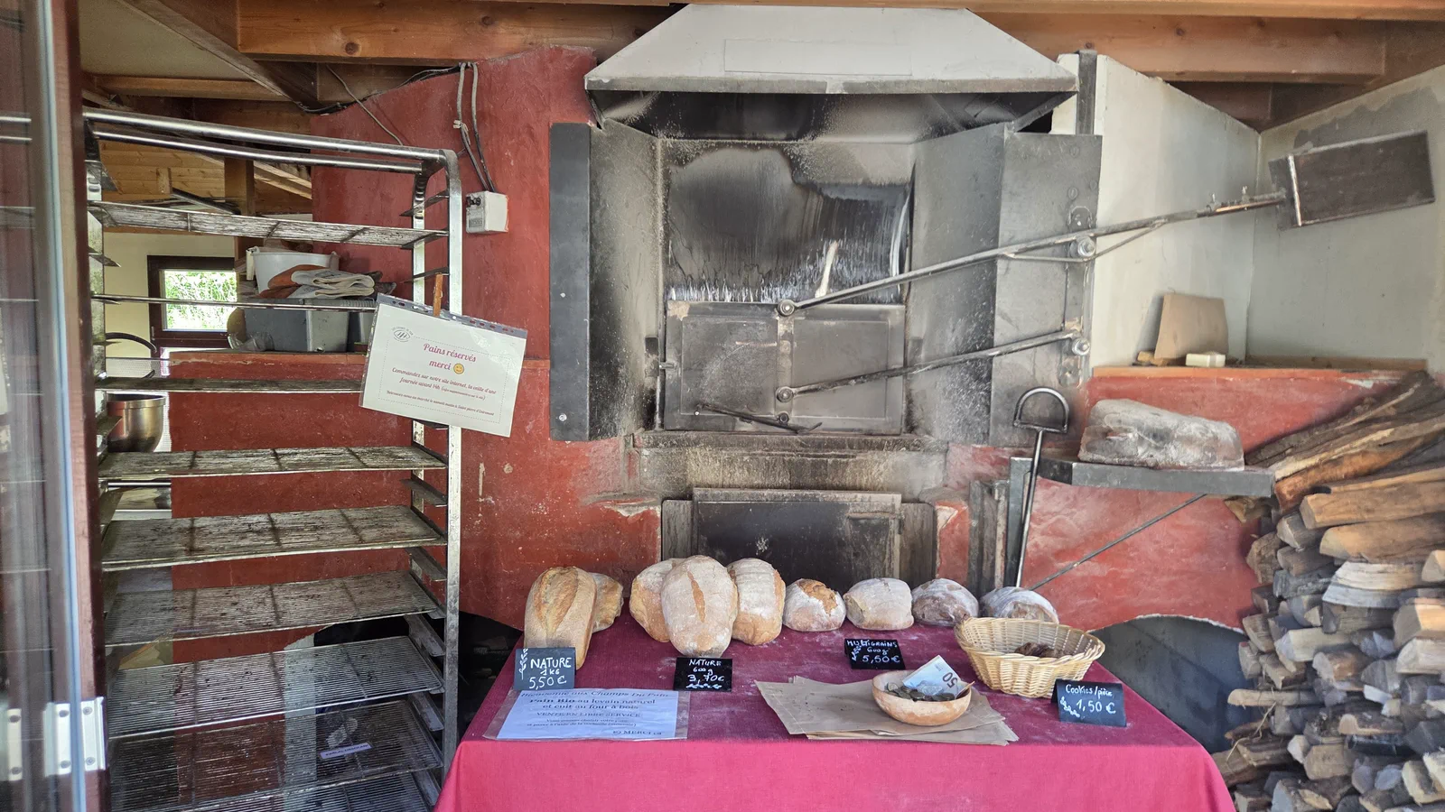 Le four à bois maçonné, peint en rouge et noirci par la flamme, avec devant une table nappée de rose où sont alignés plusieurs pains au levain et des ardoises de prix, échelle à pains et bûches sur les côtés