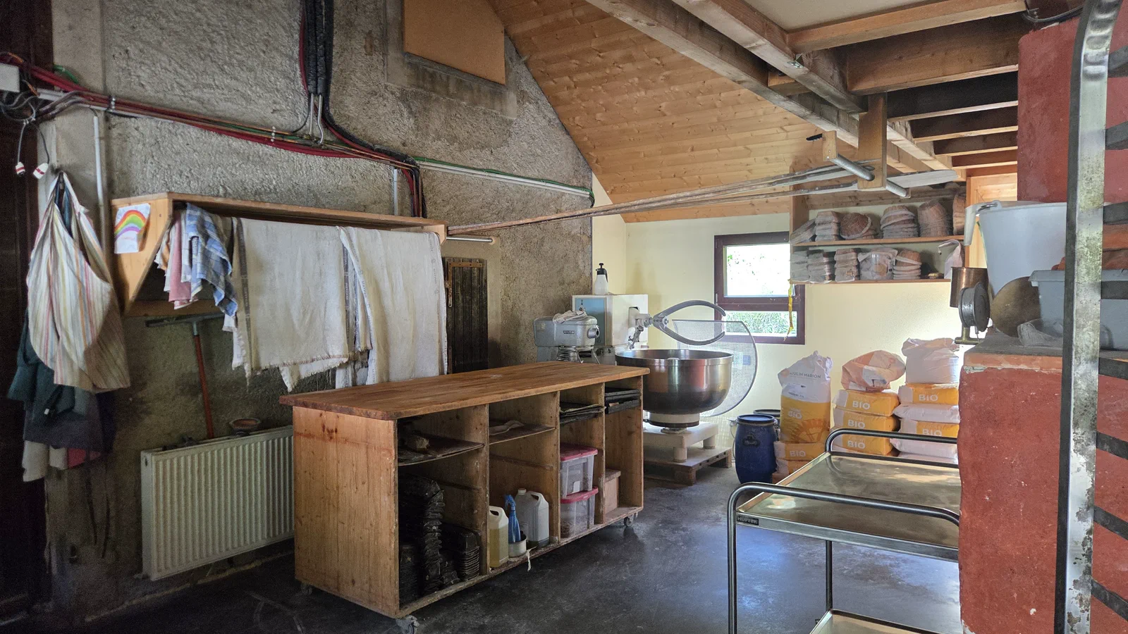 Intérieur du fournil : grand plan de travail en bois sur roulettes, pétrin et cuve inox, sacs de farine bio empilés, charpente apparente et enduit à la chaux