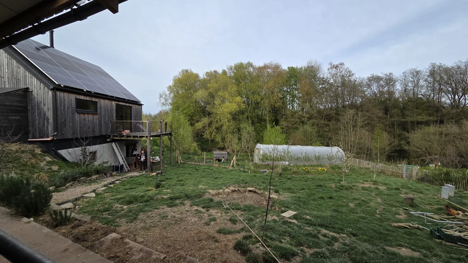 Maison bois compacte posée sur une cave apparente, panneaux photovoltaïques en toiture, tunnel maraîcher et jardin devant