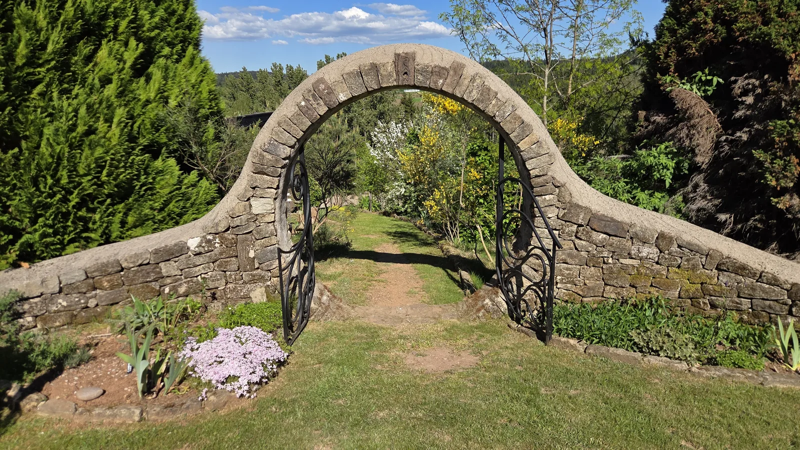 Grande arche en pierre sèche en forme de cercle, fermée par une grille en fer forgé, ouvrant sur une allée du jardin bordée de fleurs, collines boisées au fond