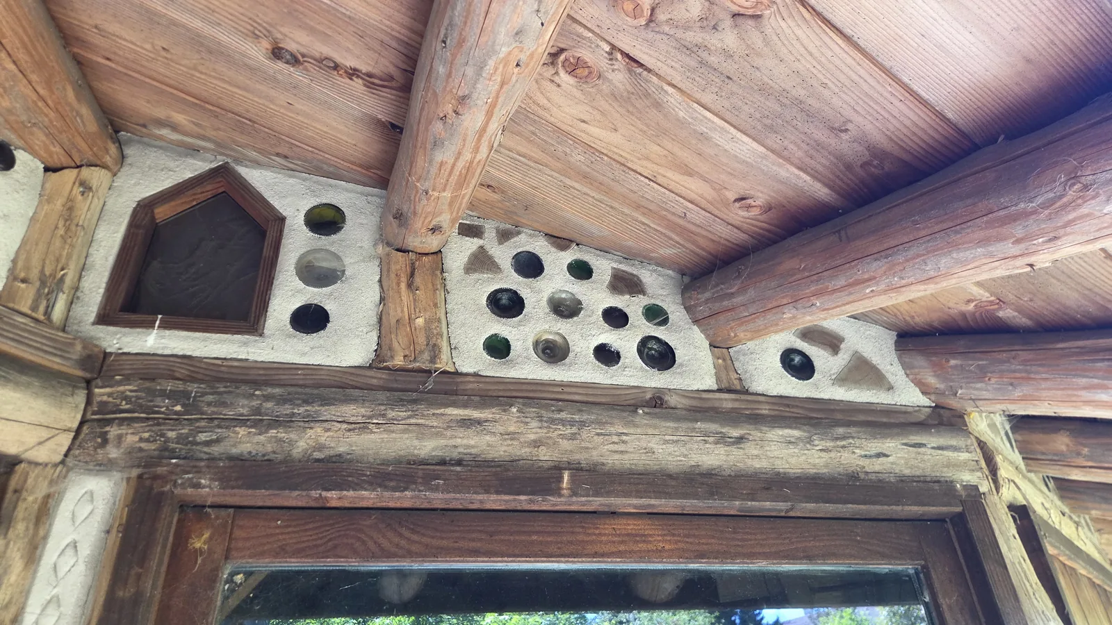 Détail d'un mur en torchis serti de culs de bouteilles en verre colorés qui laissent passer la lumière, charpente et poutres en bois autour