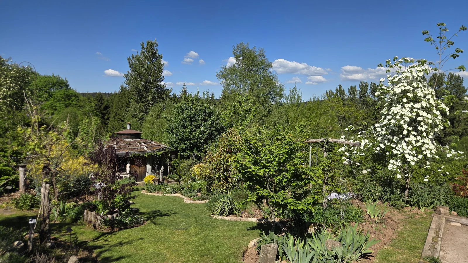 Vue d'ensemble du jardin : pelouse centrale, massifs fleuris, cabane en bois à gauche, arbustes et arbres variés, collines boisées et ciel bleu au fond