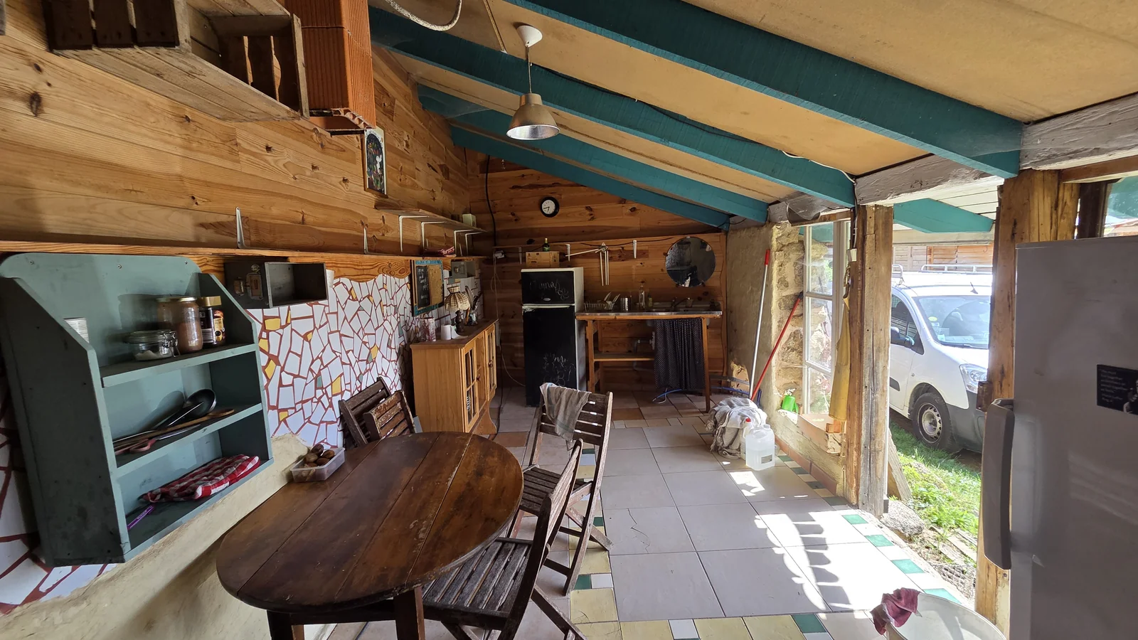 Cuisine extérieure couverte avec table et chaises en bois, étagères, mosaïque murale et un van garé juste à côté