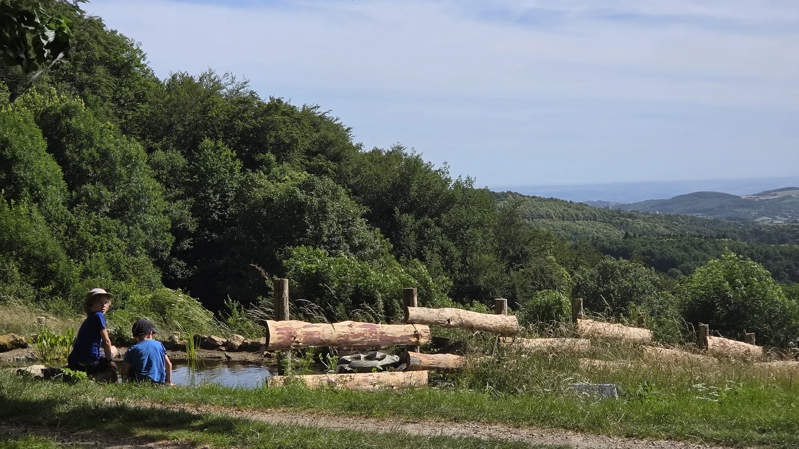 Deux enfants assis au bord d'une petite mare bordée de rondins, avec une vue dégagée sur les collines boisées de l'Ardèche