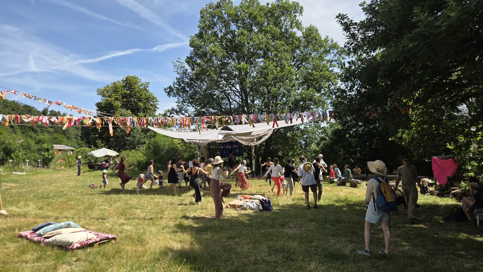Groupe de personnes dansant en cercle dans une prairie sous de grandes voiles blanches tendues et des guirlandes, lors du festival