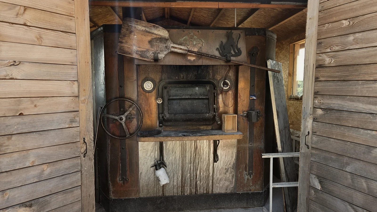 Grand four à pain en bois et métal, avec sa pelle de boulanger posée en travers, dans un fournil aux murs de bois