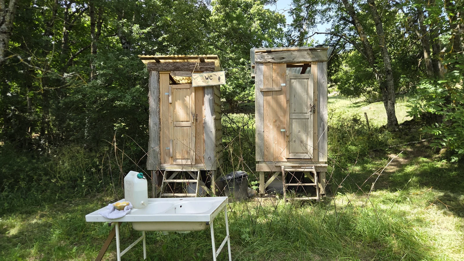 Deux cabanes de toilettes sèches en bois surélevées dans l'herbe, avec une table et un bidon pour se laver les mains devant