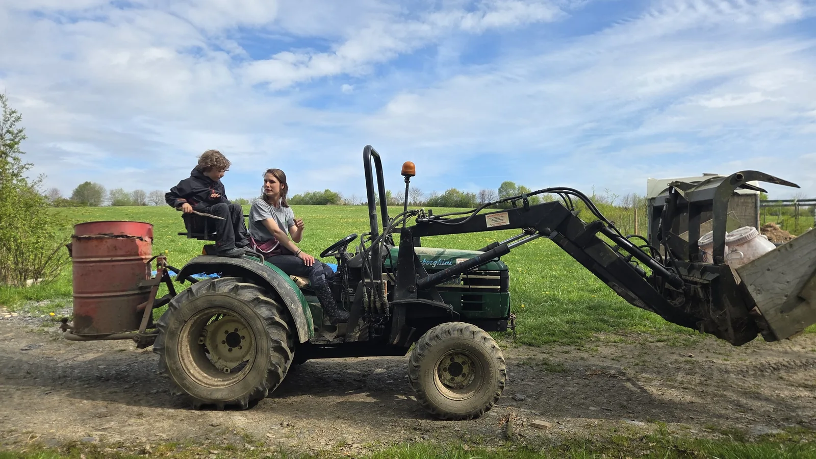 Léon assis sur le siège passager du tracteur, Justine au volant, une benne pleine accrochée à l'avant, prairie verte et ciel nuageux derrière