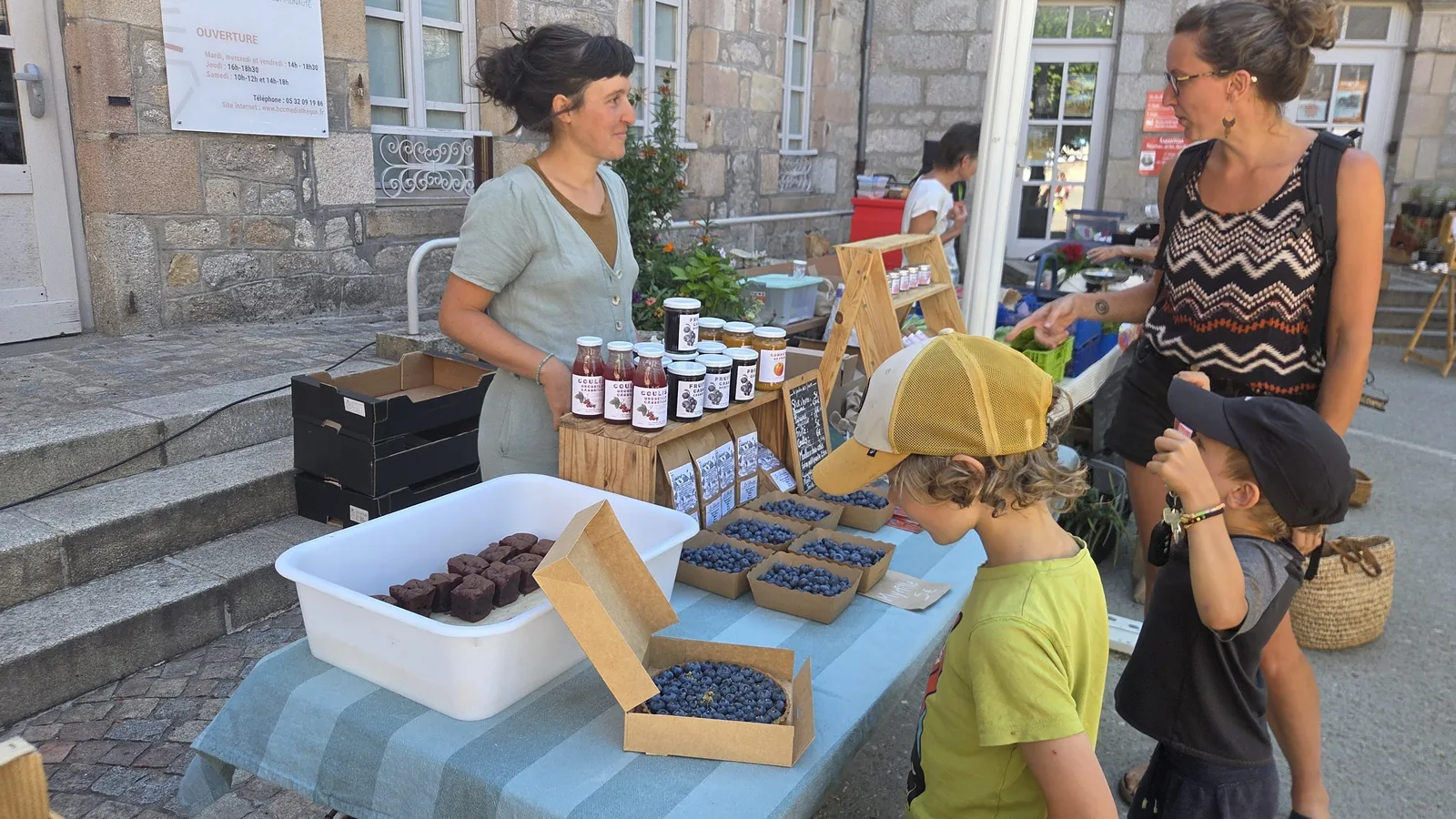 Charlotte à son stand de marché, présentant ses petits fruits et ses produits transformés, des enfants au premier plan