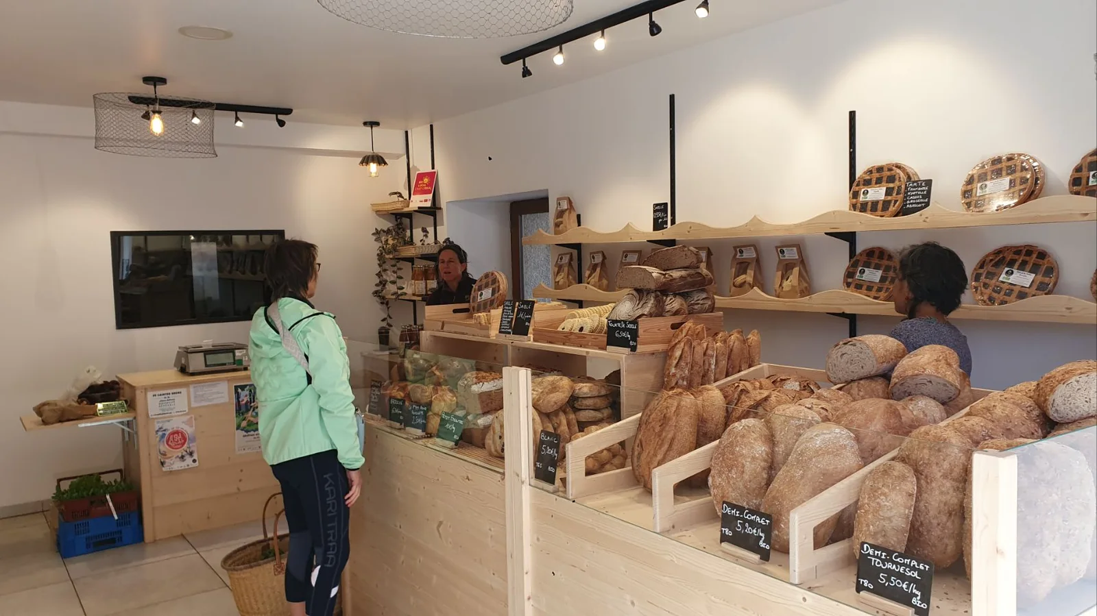 Intérieur du magasin côté boulangerie : étagères de pains au levain et de tartes, une cliente devant le comptoir en bois