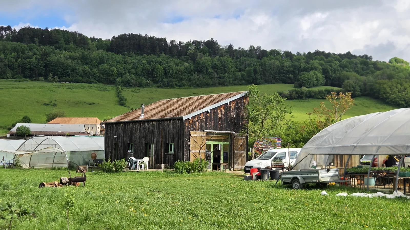 Vue de la ferme depuis le pré : la maison en bois noir au centre, deux serres tunnel de part et d'autre, collines boisées en arrière-plan