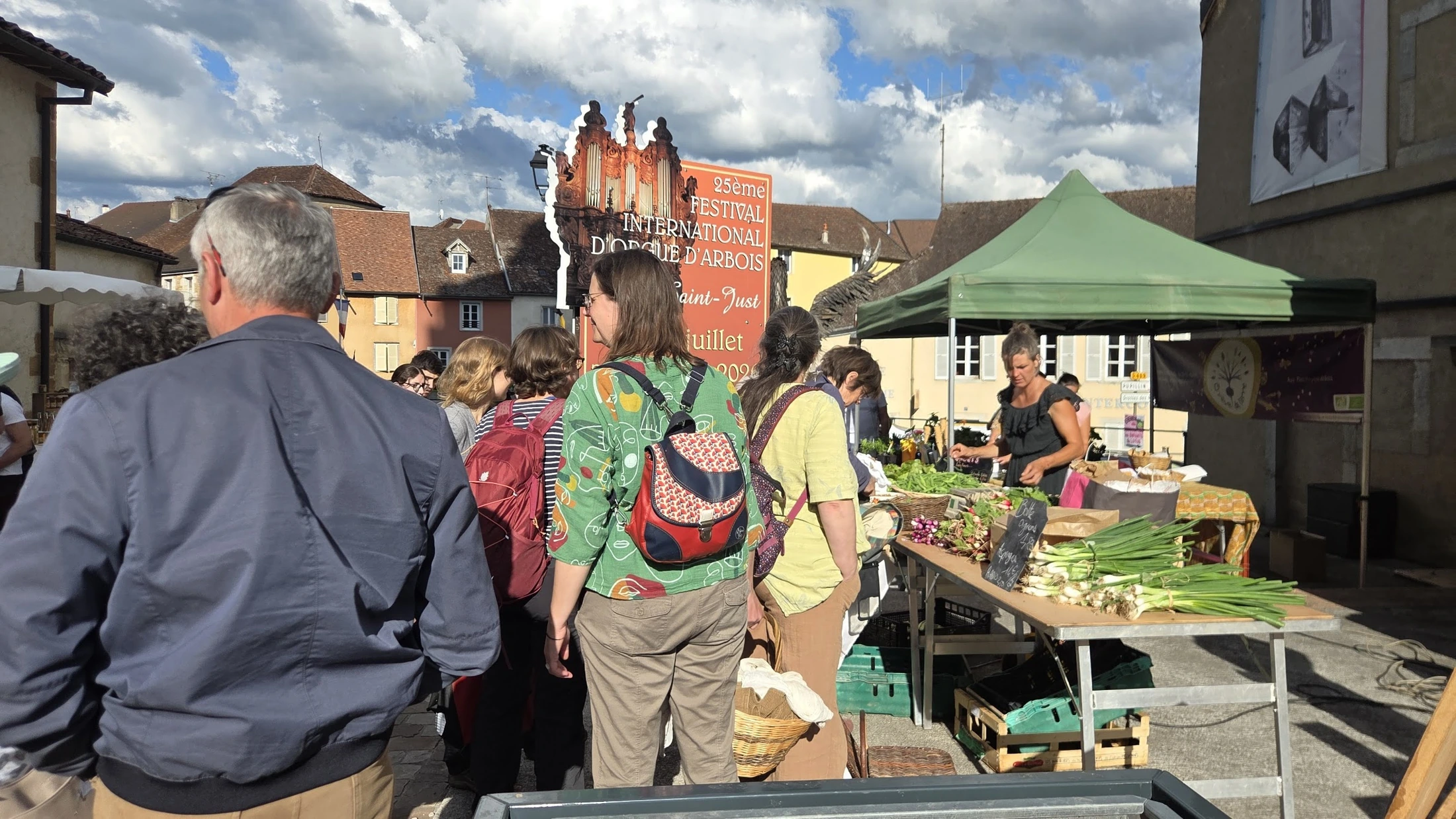 Béné dans son stand sous tente verte au marché d'Arbois, plusieurs clients qui patientent devant l'étal, panneau « Festival international d'Arbois » et bâtiments en arrière-plan